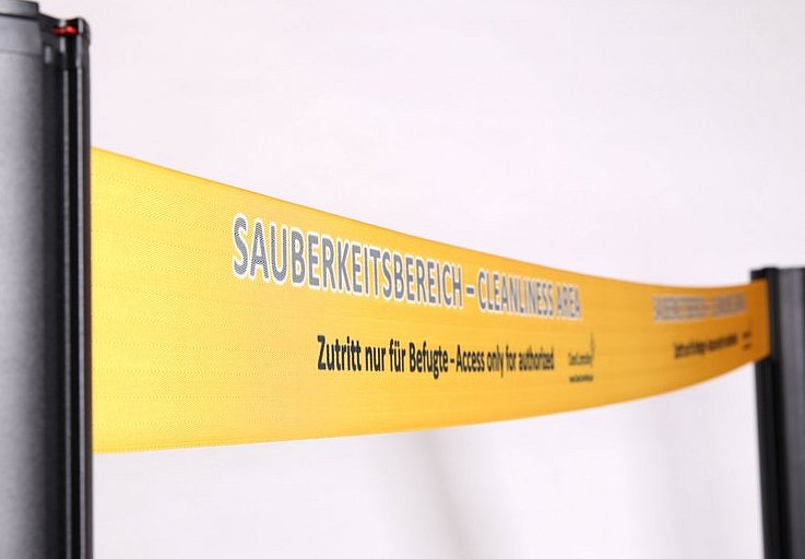 Orangenes Absperrband auf dem in blauer und schwarzer Schrift 'Sauberkeitsbereich-Cleanliness Area Zutritt nur für Befugte-Access only for athorized' steht.