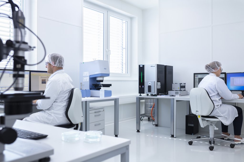 Zwei Personen, steril gekleidet sitzen vor dem Computer, neben ihnen unterschiedliche Laborinstrumente.