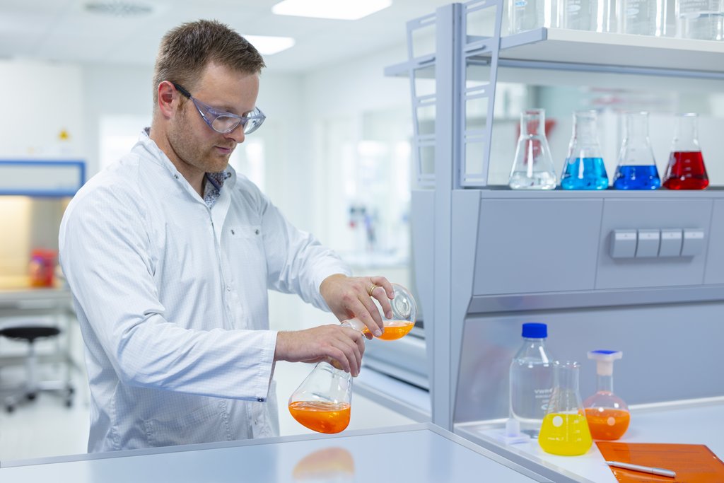 Kurzhaariger Mann mit Schutzbrille und weißen Kittel in Laborumgebung, schüttet zwei orangene Flüssigkeiten, welche sich in zwei Kolben befinden, zusammen.