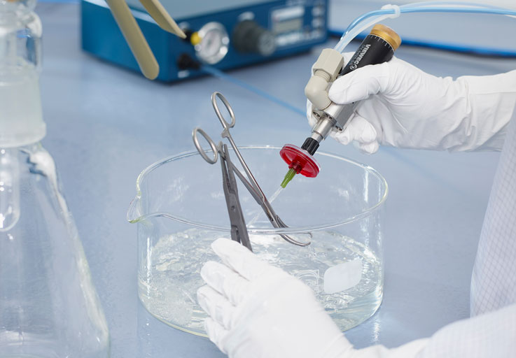 Eine große durchsichtige Schüssel mit durchsichtiger Flüssigkeit, in welcher zwei Hände in weißen Handschuhen Labor Equipment halten.